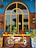 A colorful outdoor scene with a decorative bicycle frame design attached to a brick wall. The window above the bike has a bright yellow arch and is surrounded by various potted plants. The background features a wooden and blue facade, with ornate patterns on the side ceramic planters.