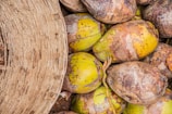 A collection of coconuts with rough, textured husks is piled closely together. On the left side of the image, a woven basket made of natural fibers is partially visible, suggesting a scene of harvesting or storage.