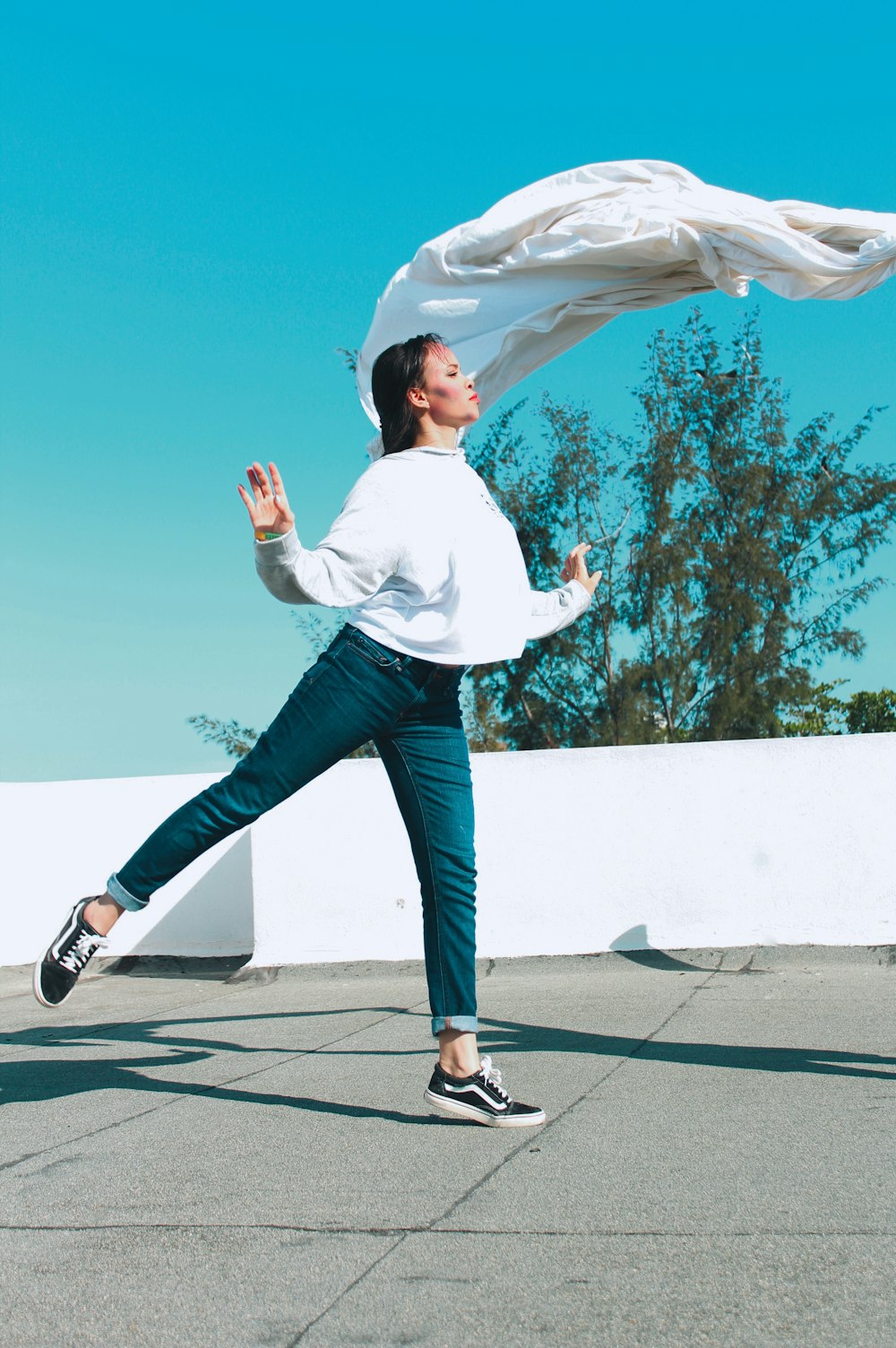 woman in white shirt and blue denim jeans jumping on gray concrete pavement during daytime