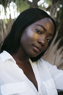 A person with long black hair is wearing a white shirt against a background of blurred greenery and branches. The lighting creates a soft, natural look with highlights on the face.