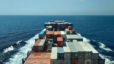 A large cargo ship loaded with containers sailing on deep blue ocean under a clear sky.