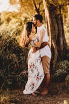 A couple embraces warmly in a lush, forested area. The woman, wearing a white floral dress, and the man, in a white shirt and khaki shorts, appear to be deeply connected with each other as he gently kisses her forehead. The background features tall trees and greenery, casting a warm, golden light.
