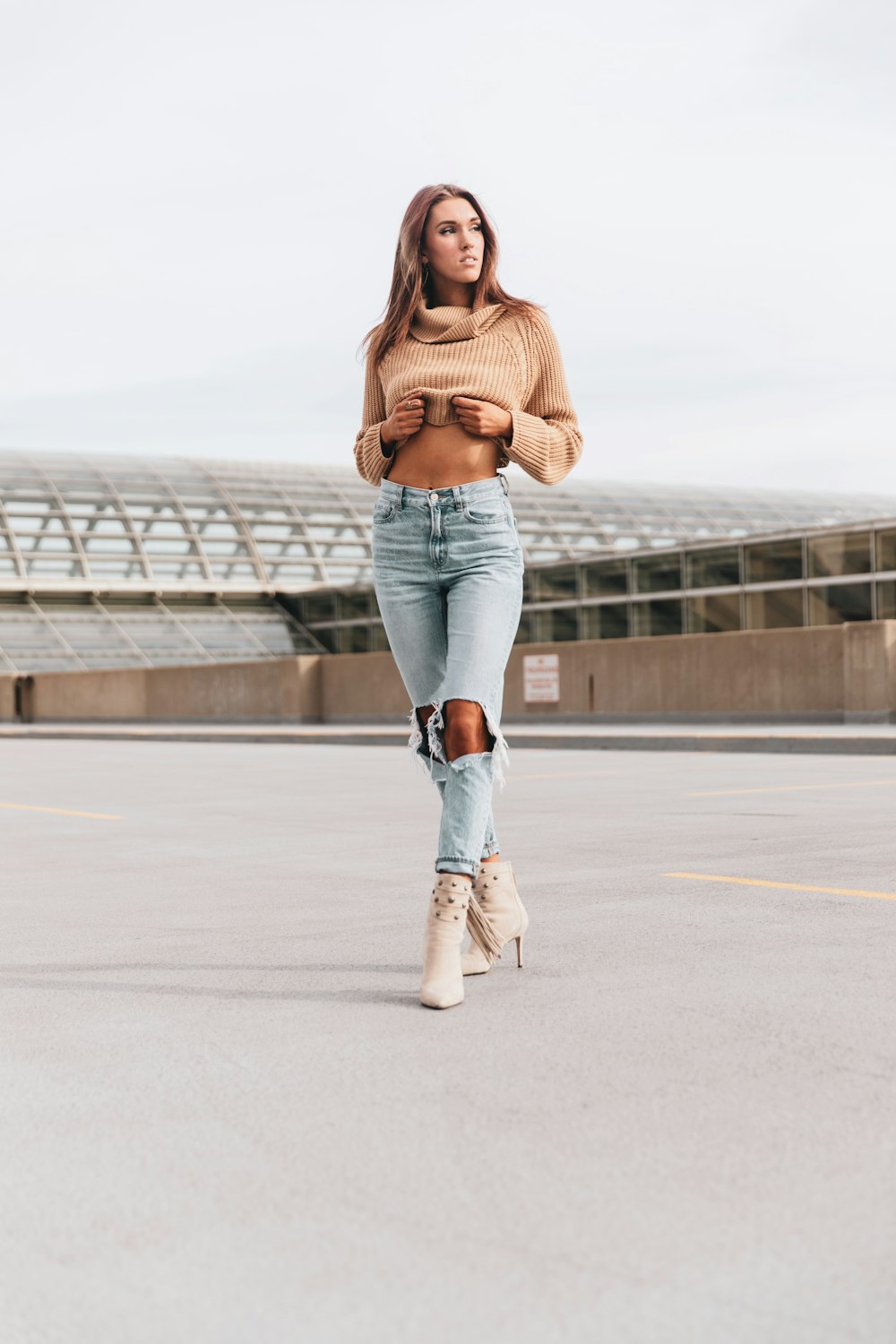 woman in brown sweater and blue denim jeans standing on gray concrete road during daytime