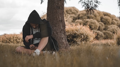 A person wearing a hoodie sits on the grass next to a tree, holding a smartphone. The individual is looking down, creating a sense of contemplation or focus. The setting is a serene natural environment with grassy fields and thick, leafy trees in the background. The overall atmosphere is calm and introspective.