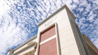 A large, modern building viewed from a low angle with a detailed architectural design and a facade featuring red brick inlays. The building's name and possibly departments are visible, located on an ornate part of the structure. In the background, a sky filled with scattered clouds complements the building's exterior.