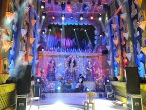 A vibrant stage setting features elaborate sculptures and decorations, dominated by figures of deities in traditional attire. The backdrop is richly adorned with intricate designs and colorful lighting, creating a festive ambiance.