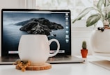 A minimalist white mug resting on a wooden desk next to a laptop and a small potted plant.