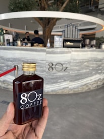 A hand holding a bottle labeled '80z Coffee' in front of a modern coffee counter with a barista and coffee-making equipment. The counter has a sleek, marble-like appearance with a tree integrated into the design.
