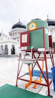 A handwashing station is set up in a mosque courtyard featuring a large green water tank mounted on a red metal frame. The background shows the mosque’s iconic architecture with domed roofs and white walls.