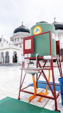 A handwashing station is set up in a mosque courtyard featuring a large green water tank mounted on a red metal frame. The background shows the mosque’s iconic architecture with domed roofs and white walls.