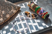 A woven pillow lies on a textured surface next to a pair of sunglasses and a colorful cylindrical case with intricate patterns. Sunlight creates dappled shadows across the scene.
