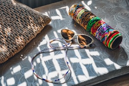 A woven pillow lies on a textured surface next to a pair of sunglasses and a colorful cylindrical case with intricate patterns. Sunlight creates dappled shadows across the scene.
