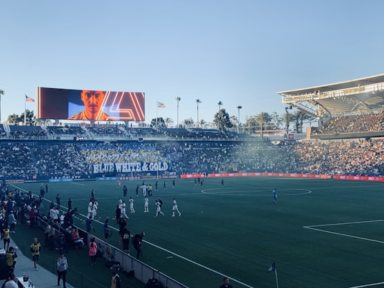 A large sports stadium filled with spectators. The field is occupied by soccer players, some walking and others scattered across the pitch. A massive screen displays a person, and flags fly on top of the stadium roof. The audience is dense, and the atmosphere seems lively with a visible banner showing 'BLUE WHITE & GOLD'.