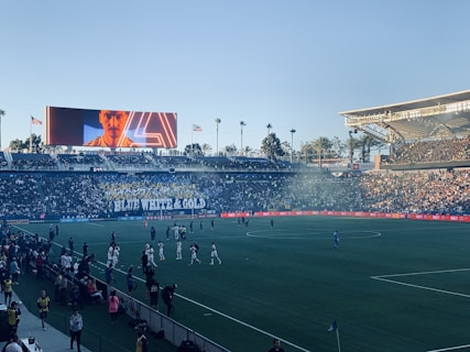 A large sports stadium filled with spectators. The field is occupied by soccer players, some walking and others scattered across the pitch. A massive screen displays a person, and flags fly on top of the stadium roof. The audience is dense, and the atmosphere seems lively with a visible banner showing 'BLUE WHITE & GOLD'.