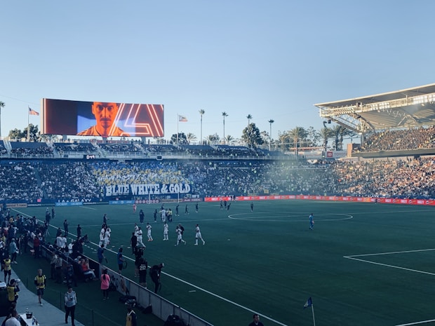 A large sports stadium filled with spectators. The field is occupied by soccer players, some walking and others scattered across the pitch. A massive screen displays a person, and flags fly on top of the stadium roof. The audience is dense, and the atmosphere seems lively with a visible banner showing 'BLUE WHITE & GOLD'.