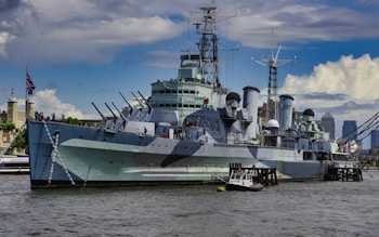 A large, historical battleship is anchored on a river with multiple turrets and radar equipment visible on deck. The ship has a camouflage pattern and is situated in an urban environment, with figures walking on the deck. A smaller boat is positioned nearby. In the background, a tower and modern cityscape are visible, partially covered by scattered clouds.