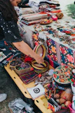 A vibrant market stall displays a variety of colorful handcrafted items, including woven baskets, embroidered textiles, and beaded jewelry. A person is interacting with the items, adding a personal touch to the lively scene. The table is covered with fabric showcasing intricate patterns and motifs, indicative of traditional craftsmanship.
