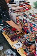 A vibrant market stall displays a variety of colorful handcrafted items, including woven baskets, embroidered textiles, and beaded jewelry. A person is interacting with the items, adding a personal touch to the lively scene. The table is covered with fabric showcasing intricate patterns and motifs, indicative of traditional craftsmanship.