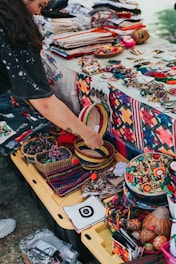 A vibrant market stall displays a variety of colorful handcrafted items, including woven baskets, embroidered textiles, and beaded jewelry. A person is interacting with the items, adding a personal touch to the lively scene. The table is covered with fabric showcasing intricate patterns and motifs, indicative of traditional craftsmanship.