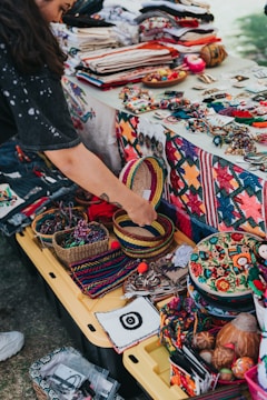 A vibrant market stall displays a variety of colorful handcrafted items, including woven baskets, embroidered textiles, and beaded jewelry. A person is interacting with the items, adding a personal touch to the lively scene. The table is covered with fabric showcasing intricate patterns and motifs, indicative of traditional craftsmanship.