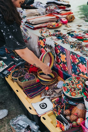 A vibrant market stall displays a variety of colorful handcrafted items, including woven baskets, embroidered textiles, and beaded jewelry. A person is interacting with the items, adding a personal touch to the lively scene. The table is covered with fabric showcasing intricate patterns and motifs, indicative of traditional craftsmanship.
