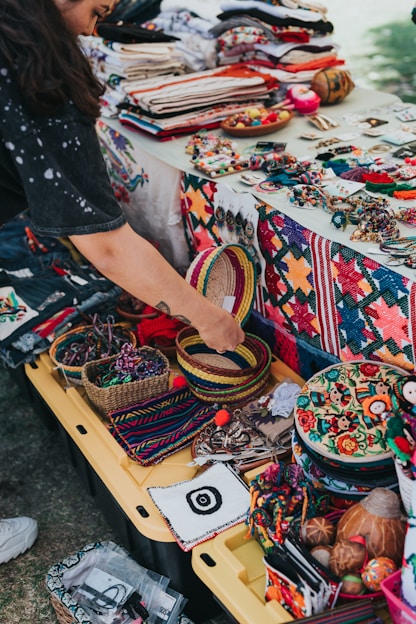 A vibrant market stall displays a variety of colorful handcrafted items, including woven baskets, embroidered textiles, and beaded jewelry. A person is interacting with the items, adding a personal touch to the lively scene. The table is covered with fabric showcasing intricate patterns and motifs, indicative of traditional craftsmanship.