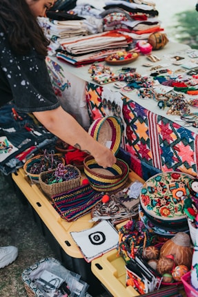 A vibrant market stall displays a variety of colorful handcrafted items, including woven baskets, embroidered textiles, and beaded jewelry. A person is interacting with the items, adding a personal touch to the lively scene. The table is covered with fabric showcasing intricate patterns and motifs, indicative of traditional craftsmanship.