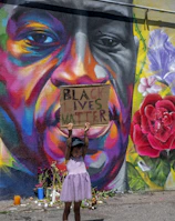 A young child wearing a purple dress and a cap holds up a sign that reads 'Black Lives Matter' in front of a colorful mural. The mural features a large face with vibrant and diverse hues such as orange, purple, and yellow. There are flowers and candles placed at the base of the mural.