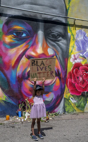 A young child wearing a purple dress and a cap holds up a sign that reads 'Black Lives Matter' in front of a colorful mural. The mural features a large face with vibrant and diverse hues such as orange, purple, and yellow. There are flowers and candles placed at the base of the mural.