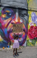 A young child wearing a purple dress and a cap holds up a sign that reads 'Black Lives Matter' in front of a colorful mural. The mural features a large face with vibrant and diverse hues such as orange, purple, and yellow. There are flowers and candles placed at the base of the mural.