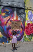 A young child wearing a purple dress and a cap holds up a sign that reads 'Black Lives Matter' in front of a colorful mural. The mural features a large face with vibrant and diverse hues such as orange, purple, and yellow. There are flowers and candles placed at the base of the mural.