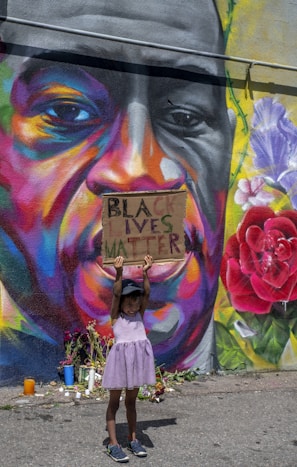 A young child wearing a purple dress and a cap holds up a sign that reads 'Black Lives Matter' in front of a colorful mural. The mural features a large face with vibrant and diverse hues such as orange, purple, and yellow. There are flowers and candles placed at the base of the mural.