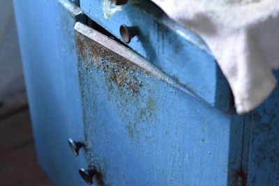 Close-up view of an old, weathered blue cabinet with visible paint chipping and rust. The cabinet has two visible handles and a white cloth draped over the top edge.