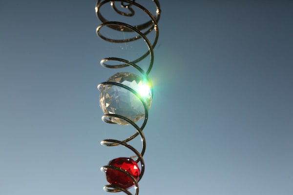 A shiny crystal and a red gem are encased in a spiral metal coil against a clear blue sky. Sunlight reflects off the crystal, creating a bright flare.