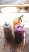 Three mason jars filled with smoothies are placed on a wooden table. The drinks have different colors and textures: one is creamy beige, another is a vibrant purple, and the third is a bright yellow with a sprig of mint as garnish. Each jar has a clear straw for drinking.