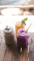 Three mason jars filled with smoothies are placed on a wooden table. The drinks have different colors and textures: one is creamy beige, another is a vibrant purple, and the third is a bright yellow with a sprig of mint as garnish. Each jar has a clear straw for drinking.