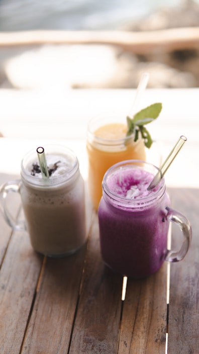 Three mason jars filled with smoothies are placed on a wooden table. The drinks have different colors and textures: one is creamy beige, another is a vibrant purple, and the third is a bright yellow with a sprig of mint as garnish. Each jar has a clear straw for drinking.