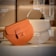 Two handbags are displayed prominently, one in a soft orange hue and the other in a light cream color. They are positioned against a minimalistic background with subtle lighting that highlights their curved shapes and clean design. Both bags have a signature clasp on the front.