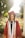 A graduate in a maroon cap and gown stands outdoors, bathed in warm sunlight with a background of blooming flowers and greenery. The individual wears honor cords and a stole, with a bright smile.