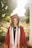 A graduate in a maroon cap and gown stands outdoors, bathed in warm sunlight with a background of blooming flowers and greenery. The individual wears honor cords and a stole, with a bright smile.