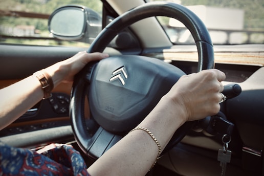 Hands are gripping a black car steering wheel with a Citroën logo visible. A side mirror reflects the outdoor environment, which appears blurred. The person is wearing a brown wristwatch on the left wrist and a gold bracelet on the right. Car keys dangle from the ignition.