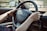 Hands are gripping a black car steering wheel with a Citroën logo visible. A side mirror reflects the outdoor environment, which appears blurred. The person is wearing a brown wristwatch on the left wrist and a gold bracelet on the right. Car keys dangle from the ignition.