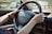 Hands are gripping a black car steering wheel with a Citroën logo visible. A side mirror reflects the outdoor environment, which appears blurred. The person is wearing a brown wristwatch on the left wrist and a gold bracelet on the right. Car keys dangle from the ignition.