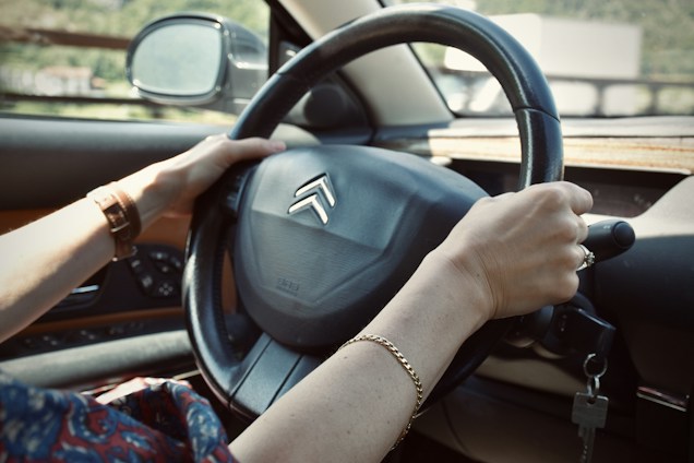 Hands are gripping a black car steering wheel with a Citro&euml;n logo visible. A side mirror reflects the outdoor environment, which appears blurred. The person is wearing a brown wristwatch on the left wrist and a gold bracelet on the right. Car keys dangle from the ignition.