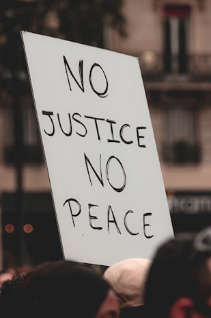 A protest sign with the words 'NO JUSTICE NO PEACE' is held above a crowd. The text is written in bold, black letters on a white rectangular board. The background shows blurred buildings and part of the crowd, creating a sense of a public demonstration.