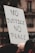 A protest sign with the words 'NO JUSTICE NO PEACE' is held above a crowd. The text is written in bold, black letters on a white rectangular board. The background shows blurred buildings and part of the crowd, creating a sense of a public demonstration.