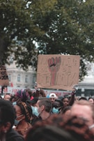 A group of people gathers in an outdoor setting with several holding signs. Most individuals are wearing masks, and some are dressed in colorful attire. Prominent in the crowd is a sign with text in French, suggesting messages related to justice and peace.