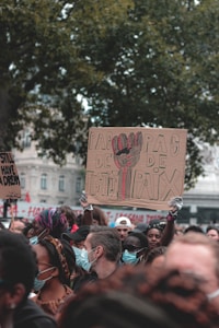 A group of people gathers in an outdoor setting with several holding signs. Most individuals are wearing masks, and some are dressed in colorful attire. Prominent in the crowd is a sign with text in French, suggesting messages related to justice and peace.