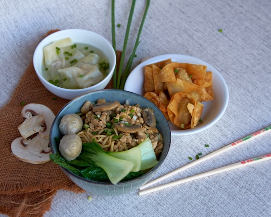 A variety of Asian dishes are arranged on a textured gray surface. In the foreground, a bowl contains noodles topped with minced meat, mushrooms, and vegetables such as bok choy and green onions. Beside it is a small white dish filled with crispy fried wontons, and another bowl with wontons in clear broth garnished with chopped chives. Fresh mushrooms, green onions, and chopsticks add to the arrangement.