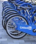 A row of blue bicycles is aligned on a tiled surface, each bicycle featuring the LinkedIn logo prominently displayed on the side panels. The bicycles are neatly arranged, suggesting they are part of a bike-sharing program or rental service.